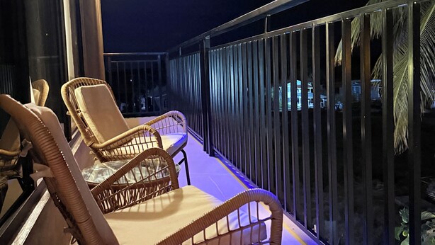 A balcony with wicker chairs and a railing, overlooking a palm tree and a building. - Jagdesh Apartments  (Anna Regina)