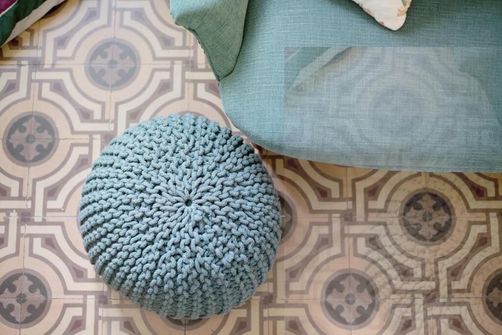 A round, knitted ottoman in a light blue color on a patterned floor.