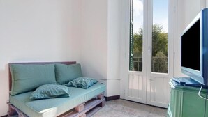 Un balcón pequeño con un colchón y almohadas, un televisor sobre un mueble bajo, y una puerta que da al exterior.