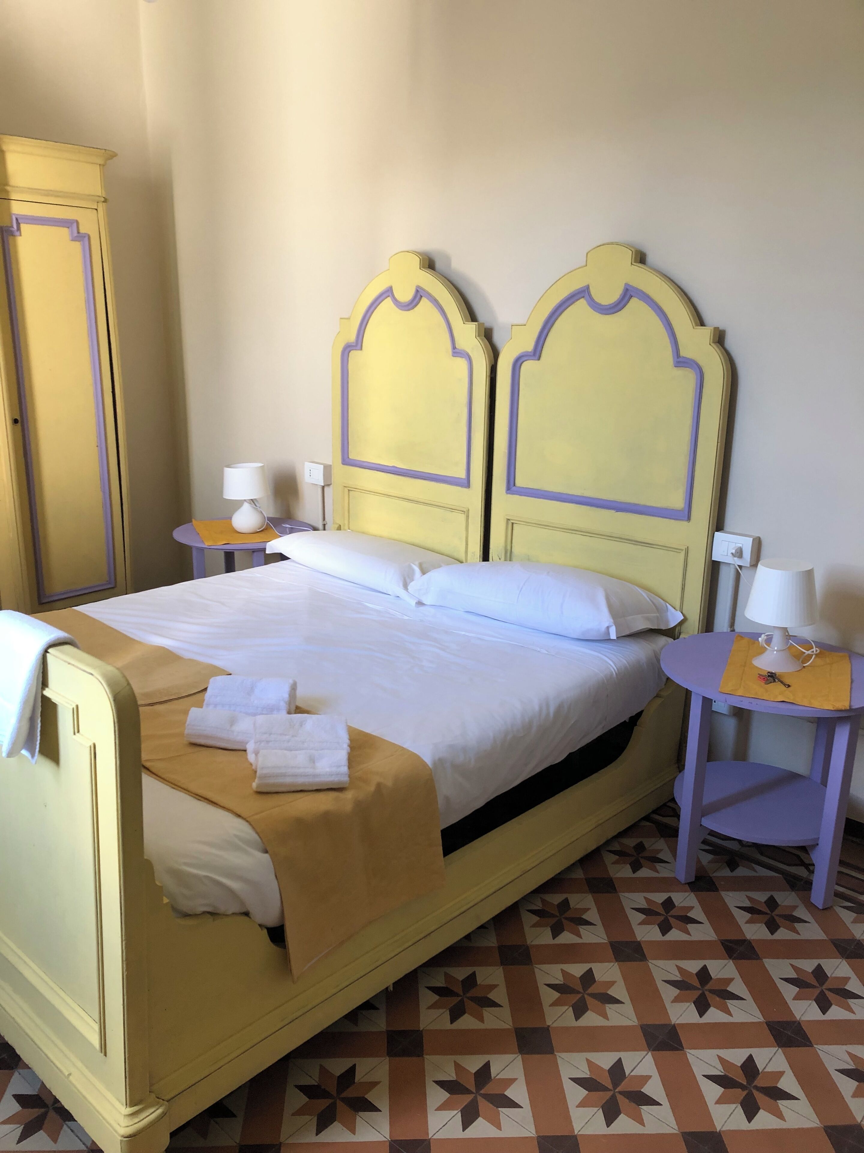 Un lit avec une tête de lit jaune et une literie blanche, une petite table ronde avec une lampe et une table de chevet assortie.