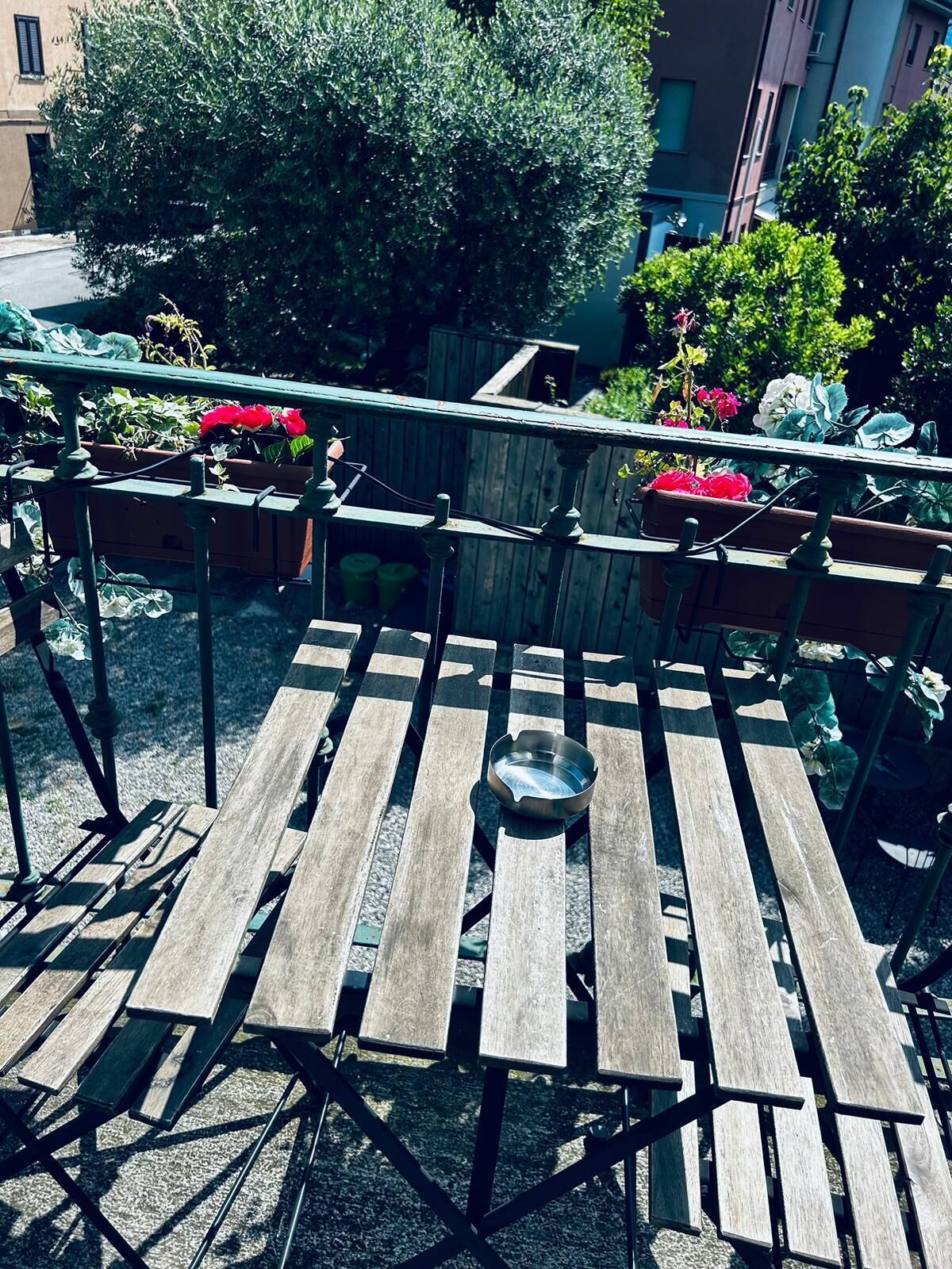 Uma área de estar ao ar livre com bancos de madeira, cadeiras de metal e uma tigela de metal sobre a mesa.