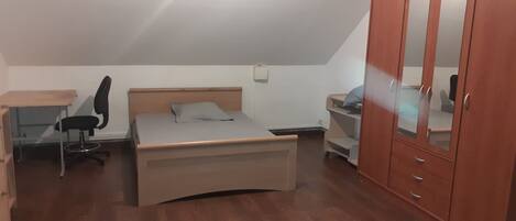 A bedroom with a bed, a desk, a chair, a wardrobe, and a small table.