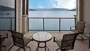A balcony with a glass table and chairs, overlooking a calm lake with mountains in the distance.