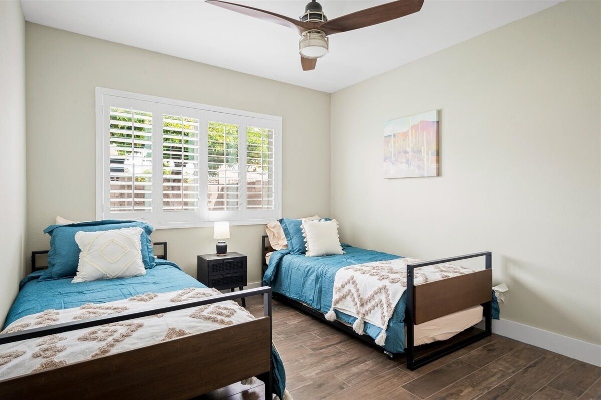 Quarto com duas camas, um ventilador de teto, janela com persianas e um quadro na parede.