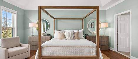 Una cama con dosel y estructura de madera, dos mesitas de noche con lámparas, una silla y un amplio ventanal con vistas.