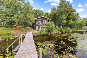 A wooden dock leading to a house with a balcony, surrounded by a pond with lily pads and lush greenery. - Cozy Rice Lake Cabin w Balcony, Grill & Views (Brainerd)