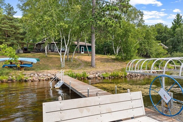 A wooden dock extending into a calm body of water, surrounded by lush greenery and a small cabin. - Cozy Rice Lake Cabin w Balcony, Grill & Views (Brainerd)