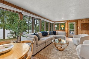 A modern living room with a large window, a sofa, a coffee table, and a dining area. - Lakefront Brainerd Cabin w Deck, Fire Pit! (Brainerd)