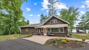 A two-story house with a covered porch, a front yard with landscaping, and a view of a lake. - Lakefront Brainerd Cabin w Deck, Fire Pit! (Brainerd)