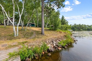 A lakeside property with a wooden deck, a pathway leading to a house, and a dock extending into the water.