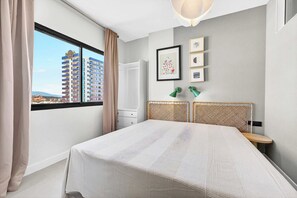 A bedroom with a large bed, a window with a view of a building, and a wall-mounted shelf with framed pictures. - Paraiso Sea Breeze Suite (Adeje)