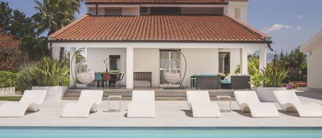 A two-story house with a swimming pool, white lounge chairs, and a patio with outdoor furniture.