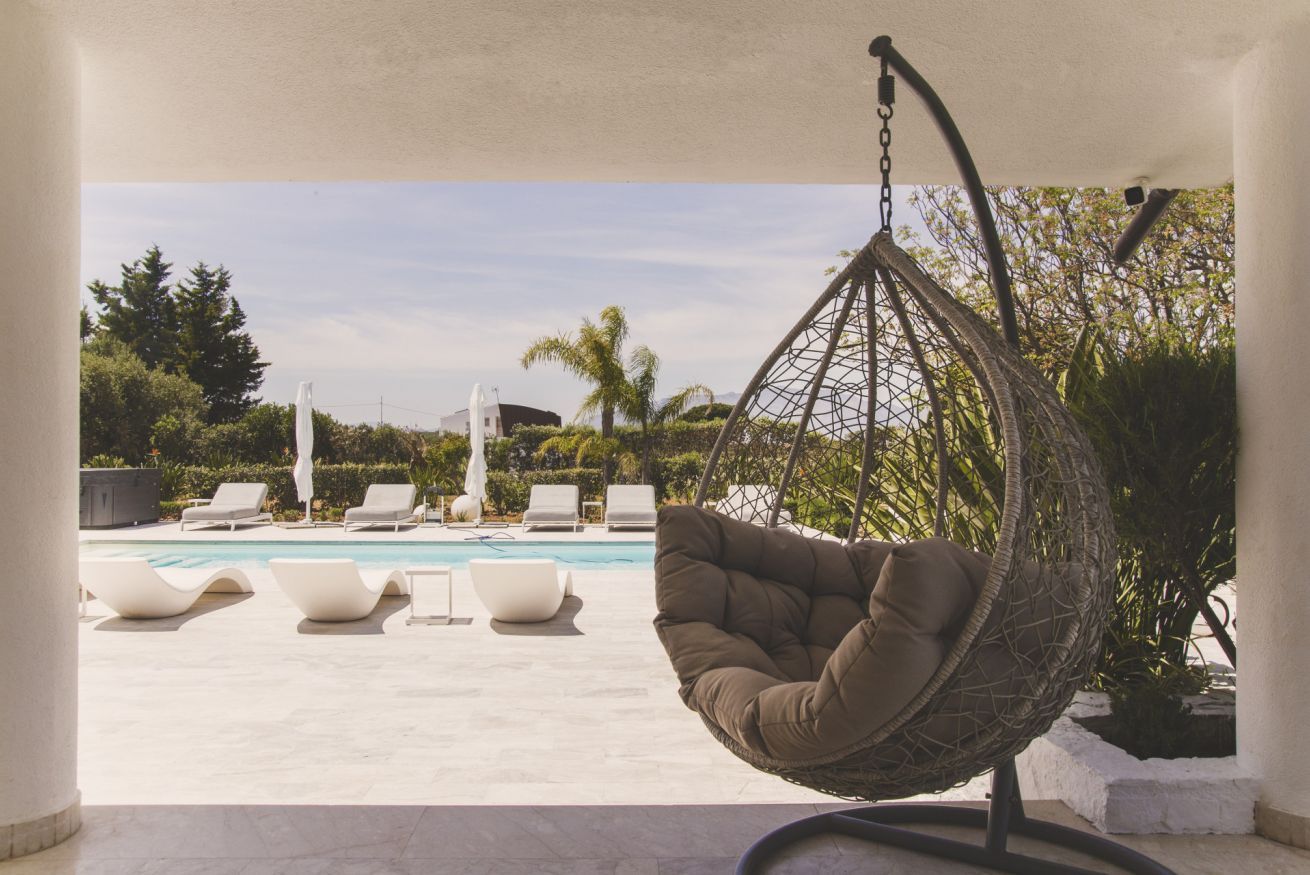 A hanging chair by a pool with lounge chairs and a view of the pool and greenery.