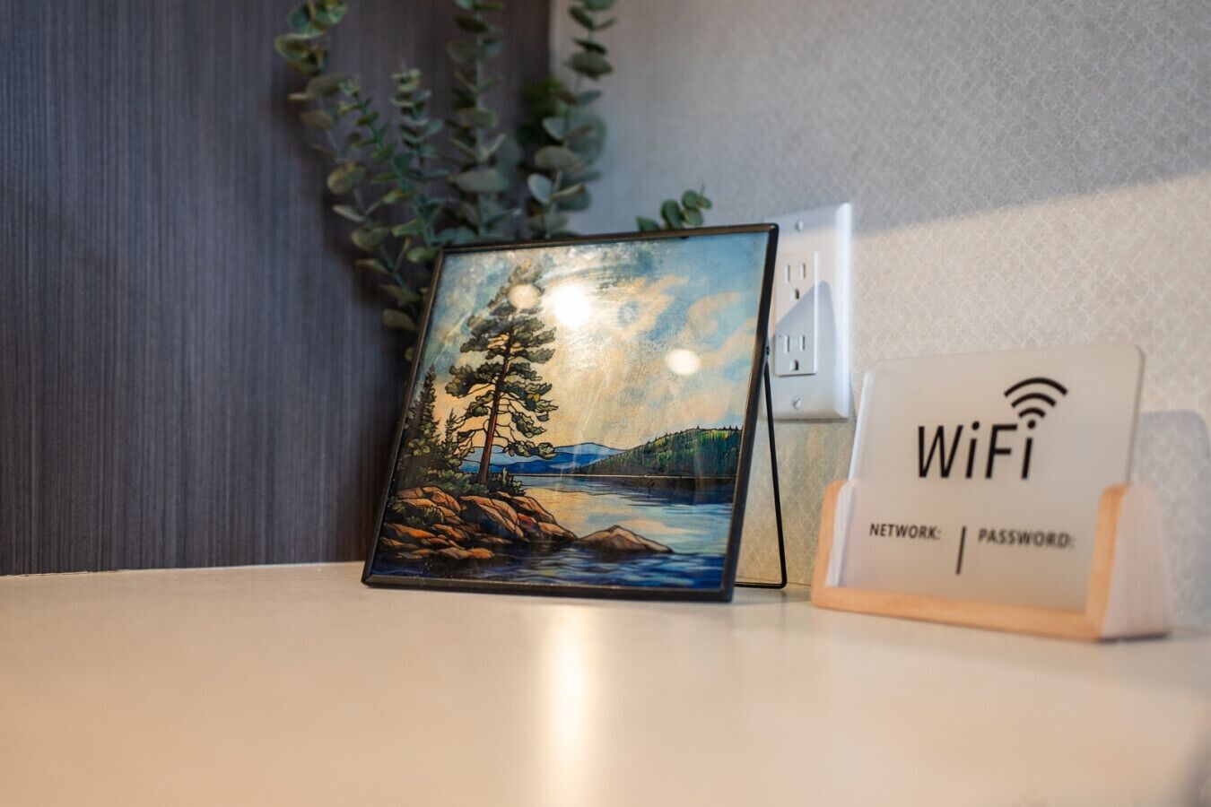 Een lijst met een schilderij van een landschap met bomen en water, een WiFi-signaal en een stopcontact op een tafel.