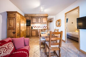 A cozy living room with a red and white checkered sofa, wooden dining table, and chairs. A kitchen with wooden cabinets and a tiled backsplash is visible in the background. - Village Lessy - Apt. O6 - Proche Station Ski (Le Grand Bornand)
