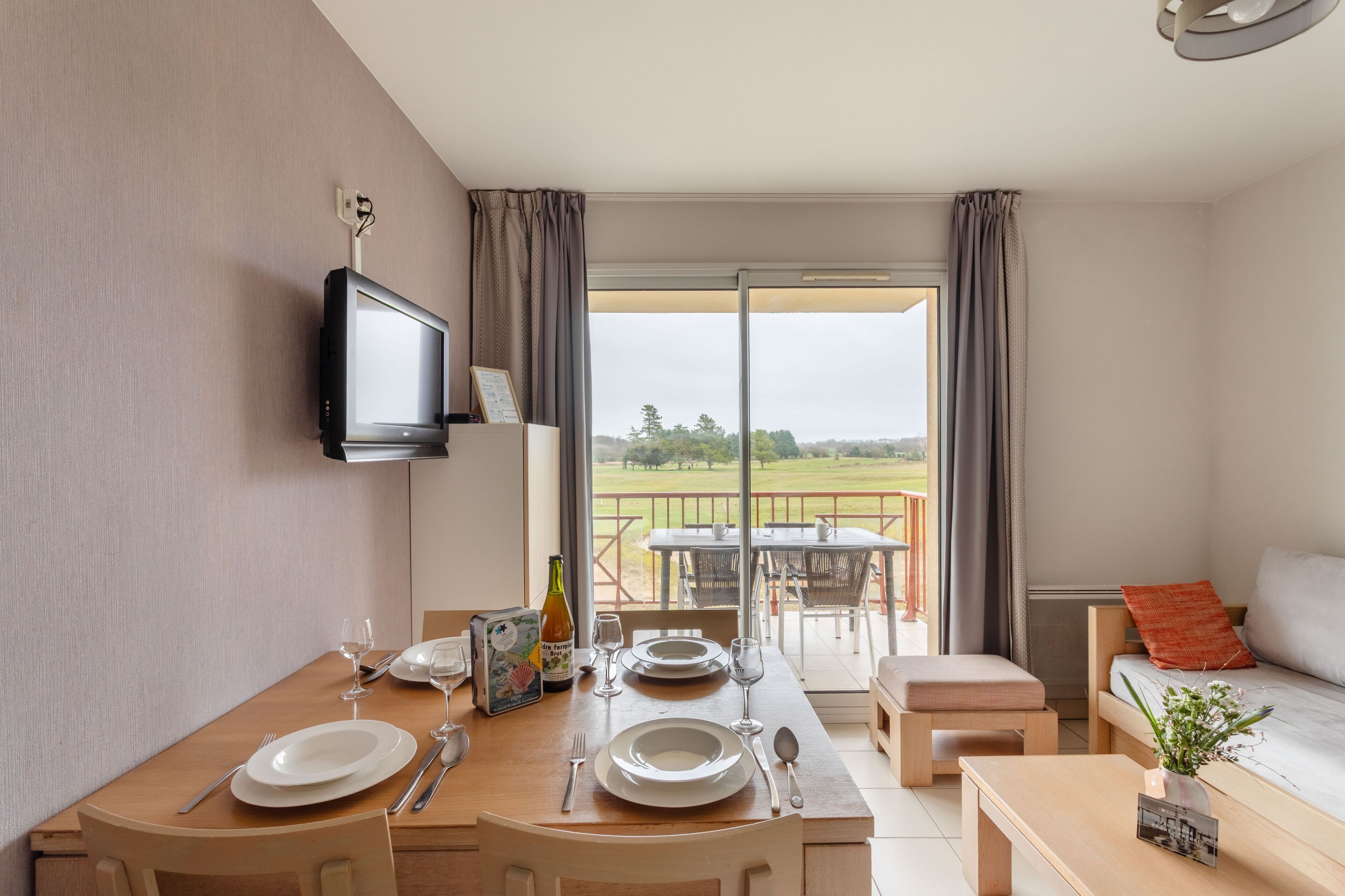 A dining area with a table set for two, a television mounted on the wall, and a view of a balcony with outdoor seating.