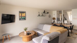 Ein modernes Wohnzimmer mit einer Couch, einem Couchtisch und einem Fernseher. Regale mit Dekorationselementen und ein Essbereich mit Tisch und Stühlen.