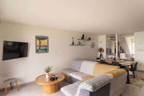 Ein modernes Wohnzimmer mit einer Couch, einem Couchtisch und einem Fernseher. Regale mit Dekorationselementen und ein Essbereich mit Tisch und Stühlen.