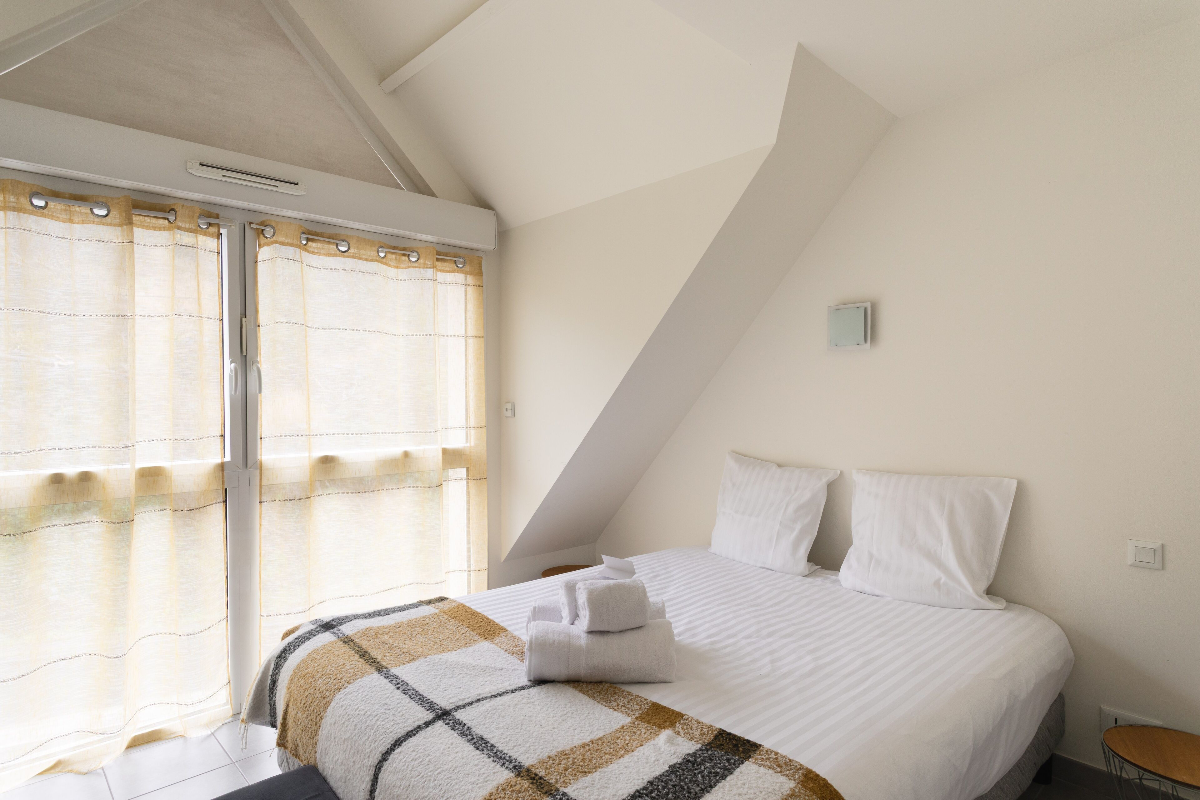 Ein Schlafzimmer mit Bett, einem Fenster mit Vorhängen, einem kleinen Tisch und einer Wandlampe.
