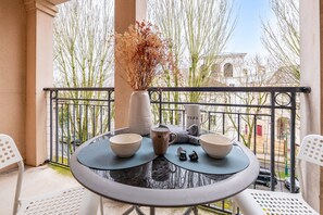 Un balcon avec une table ronde, deux bols, un vase de fleurs séchées et une vue sur des immeubles et des arbres.
