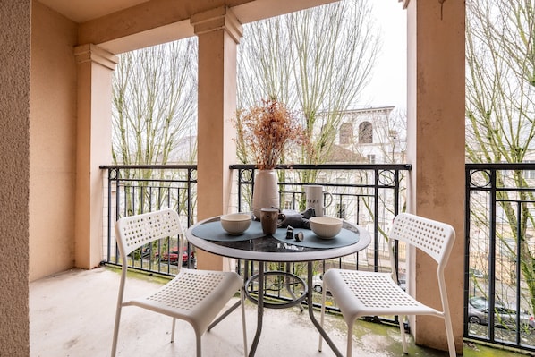 A balcony with a round table, two white chairs, a vase with dried flowers, and a view of trees and buildings.