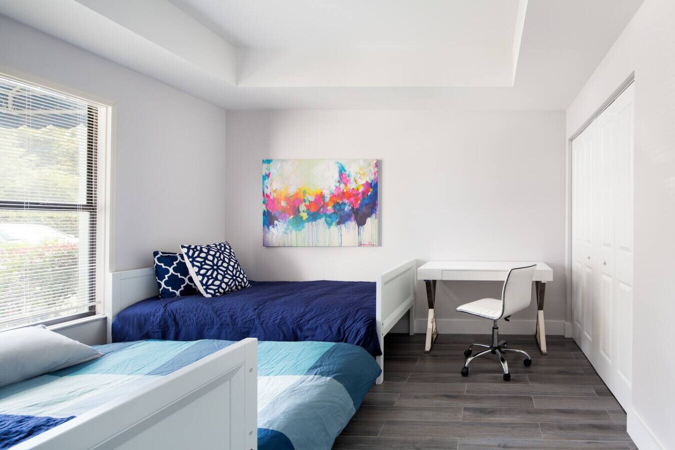 A bedroom with a bed, a desk, and a chair. There is a colorful abstract painting on the wall.