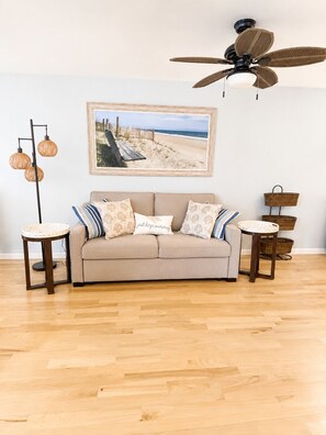Ein Wohnzimmer mit beiger Couch, gemusterten Kissen, einem gerahmten Foto vom Strand, einem Deckenventilator und Korbwäschekörben.