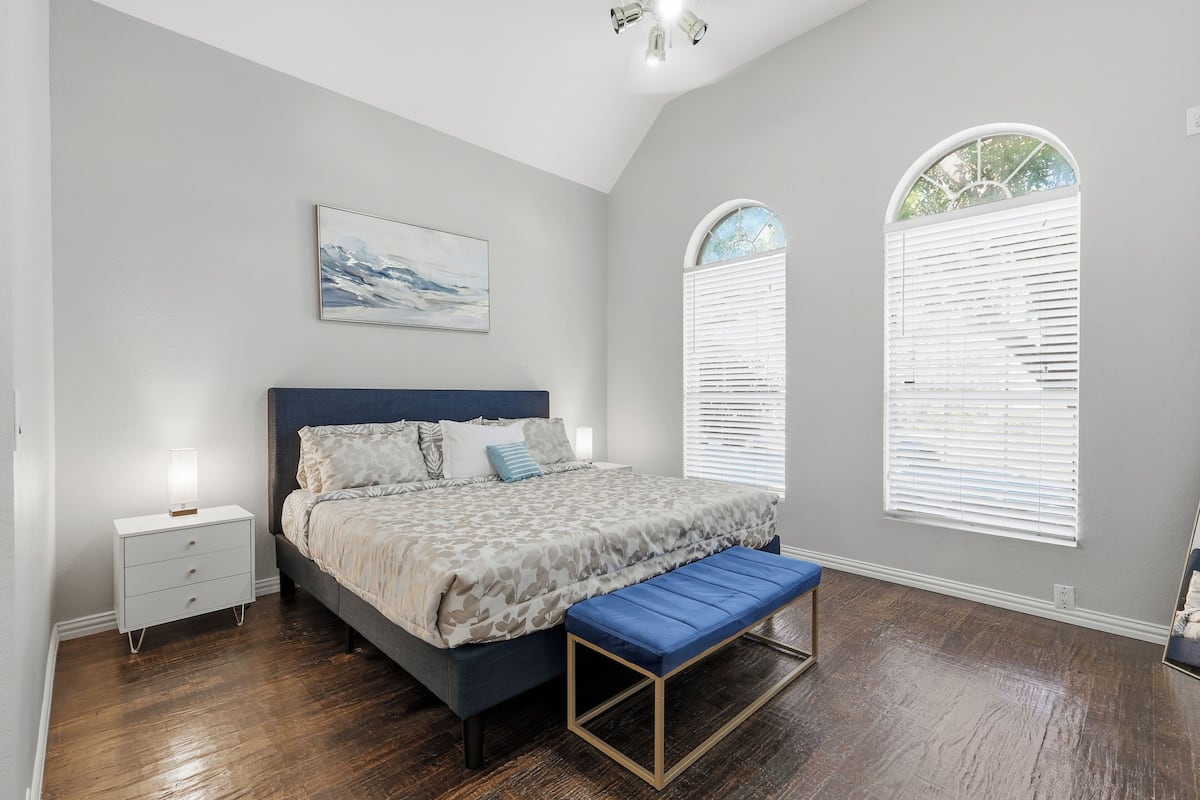 A bedroom with a bed, bedside table, bench, and two windows with blinds.