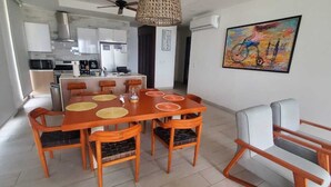Eine moderne Küche mit weißen Schränken, ein Essbereich mit einem Holztisch und orangefarbenen Stühlen sowie ein Gemälde an der Wand.