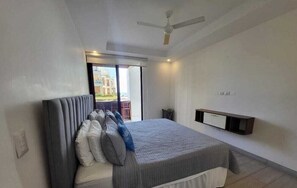 Une chambre moderne avec un grand lit, un ventilateur de plafond, une porte-fenêtre coulissante donnant sur un balcon et une étagère intégrée pour la télévision.