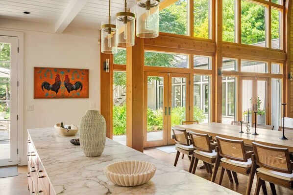 Un comedor amplio con una gran mesa de madera y sillas, una encimera de mármol y un ventanal con vistas a zonas verdes.