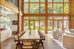 Un comedor amplio con una larga mesa de madera y sillas, ventanas grandes que ofrecen vistas al exterior y una zona de estar acogedora con sofás blancos.
