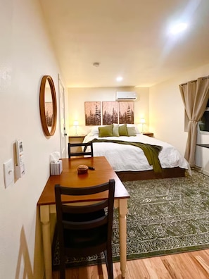 A hotel room with a bed, a table with chairs, a green patterned rug, and a wall-mounted air conditioner.