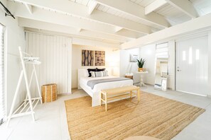 A bedroom with a bed, a wooden bench, a woven rug, a white wardrobe, and a small table with a plant.