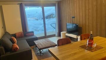 Ein Wohnzimmer mit einer Couch, einem Fernseher, einem Esstisch und Blick auf eine verschneite Landschaft durch die Glasdoor.