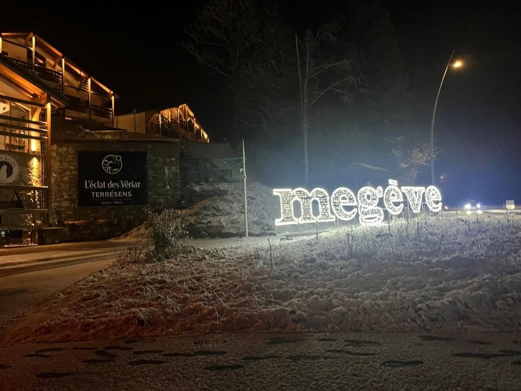 Un letrero iluminado con la palabra 'Megève', nieve, un edificio de piedra con un letrero que dice 'L'éclat des Véraz TERRESENS' y una farola al fondo.