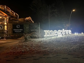 A lit sign with the word 'megève' in the snow, a stone building with a sign reading 'L'éclat des Véraz TERRESENS,' and a streetlight in the background.