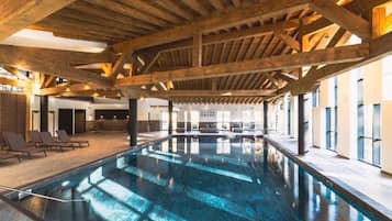 An indoor swimming pool with wooden beams, lounge chairs, and a view of the outside.