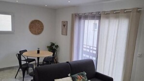 A living room with a black sofa, a round table with chairs, a small plant, and a window with curtains. - 200 m from the beach - Quiet apartment with balcony and garage num4 (Le Grau-du-Roi)