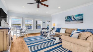 Un salon spacieux avec un canapé beige, une table basse et un tapis à rayures. On y trouve également un coin repas avec une table et des chaises, ainsi qu’un ventilateur de plafond. Un tableau représentant un voilier est accroché au mur.