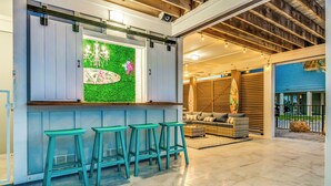Un espace bar avec des tabourets, un mur végétal agrémenté d’une planche de surf au néon, et un coin salon avec un canapé en osier et des coussins.