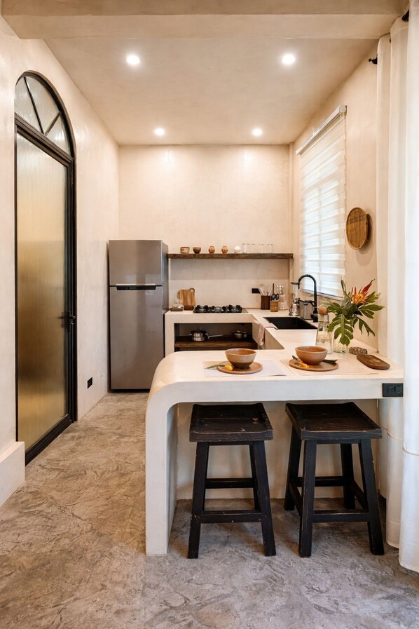 A compact kitchen with a refrigerator, stove, and sink. There are stools and a small table with bowls and utensils. - The Hidden Garden (San Marcos La Laguna)
