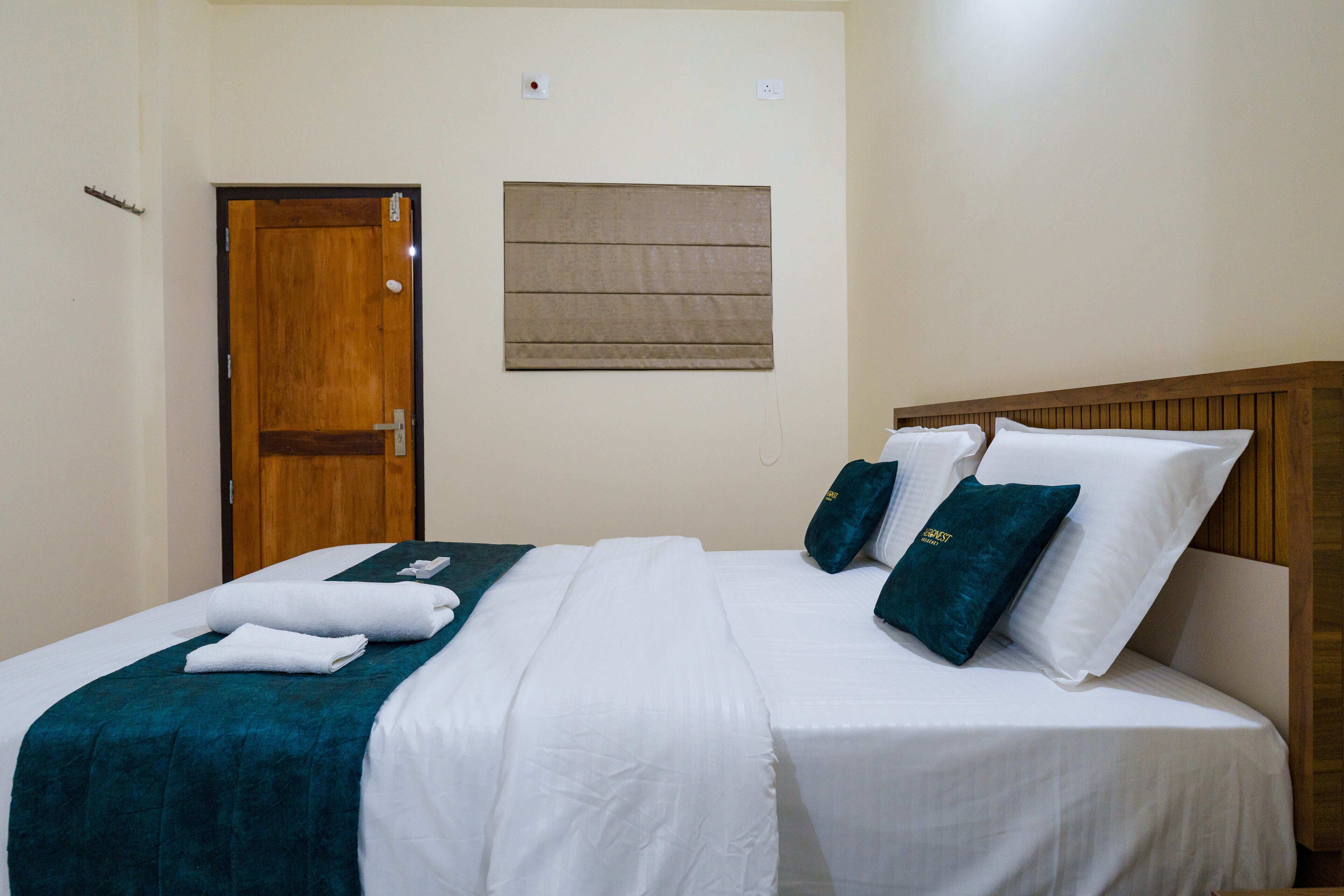 A hotel room with a wooden door, a bed with white bedding and teal accents, and a small window with a brown blind.