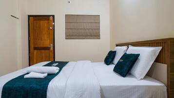 A hotel room with a wooden door, a bed with white bedding and teal accents, and a small window with a brown blind.