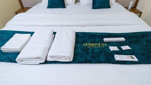 Ein ordentlich bezogenes Bett mit weißen Leinentextilien, türkisfarbenen Kissen und einer türkisfarbenen Decke mit dem Logo "Aeronest Residency". Auf dem Bett liegen zwei gefaltete weiße Handtücher und eine Fernbedienung.