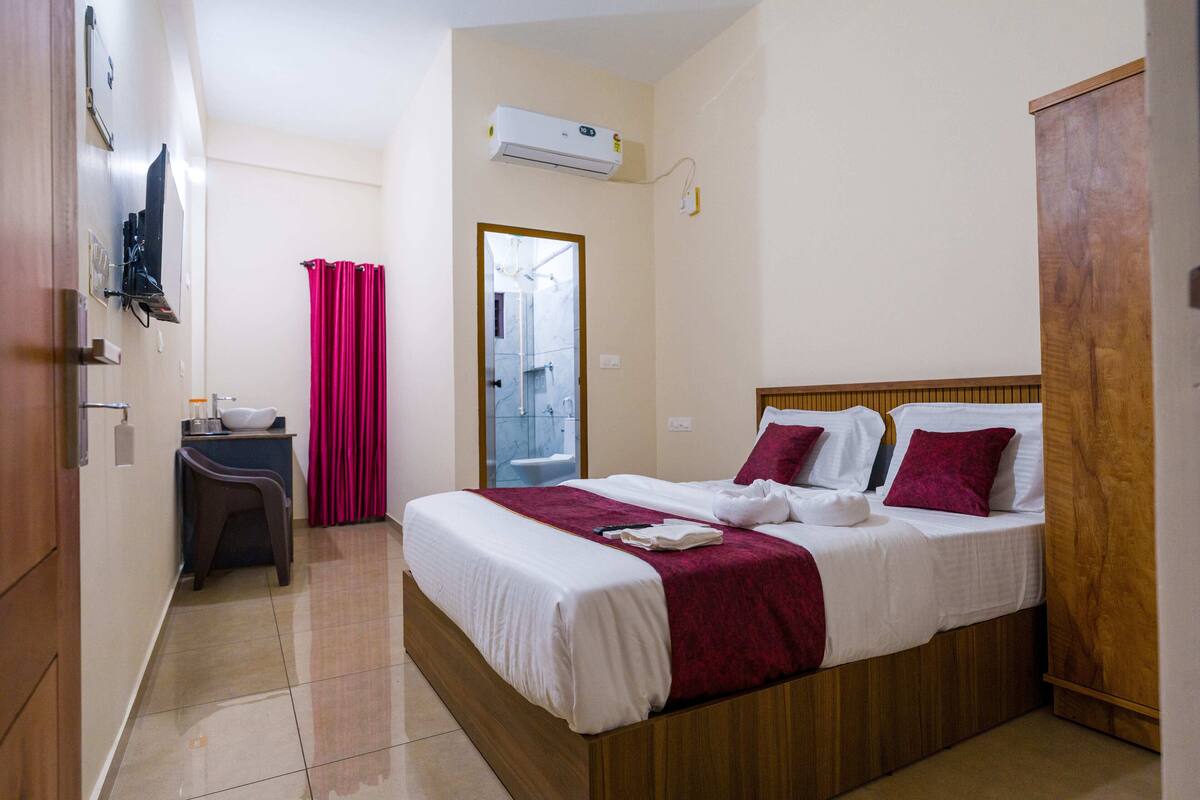 A hotel room with a wooden bed, a small desk with a chair, a bathroom visible through a glass door, and an air conditioning unit on the wall.
