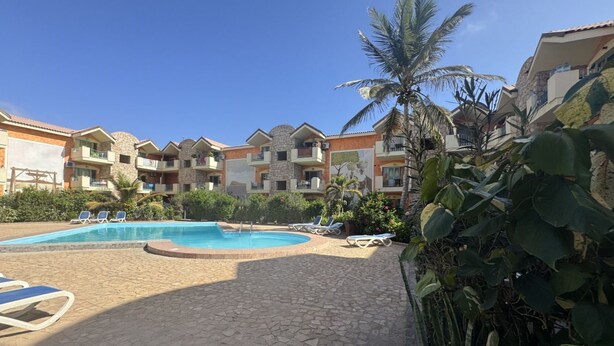 Un área de piscina con sillones de descanso, un deck de baldosas de piedra y edificios de varios pisos con balcones.