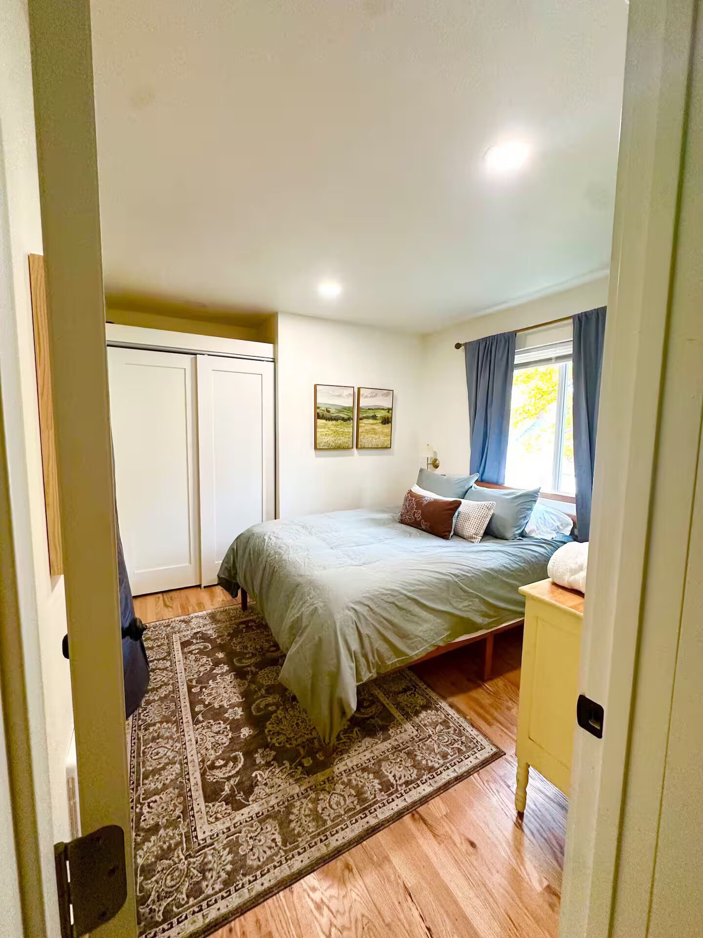 Un dormitorio con cama, mesita de noche, ventana con cortinas, puerta y pared con dos cuadros.