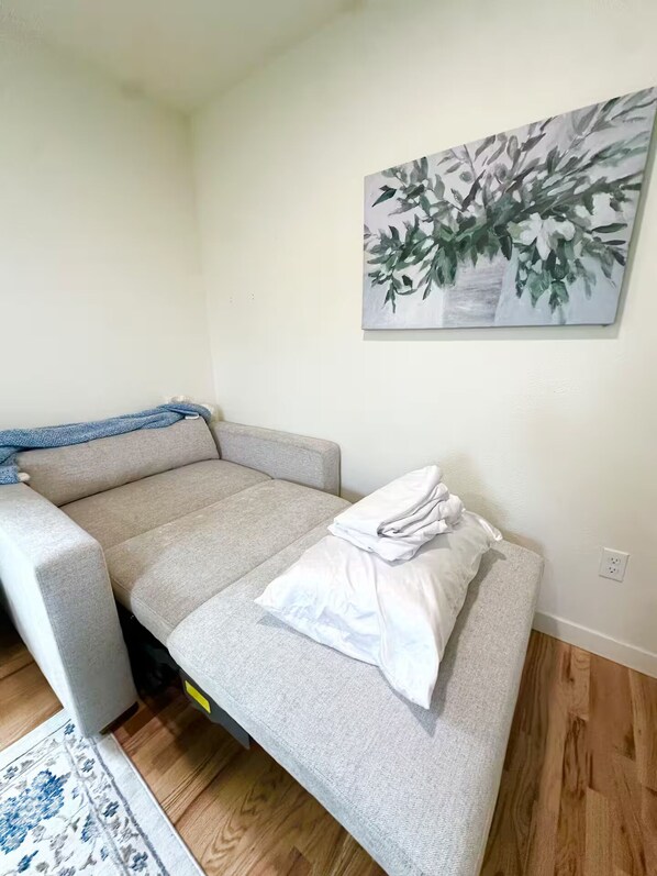 A compact room with a grey sofa, a small bed with a white pillow, a blue blanket, and a painting of leaves on the wall.