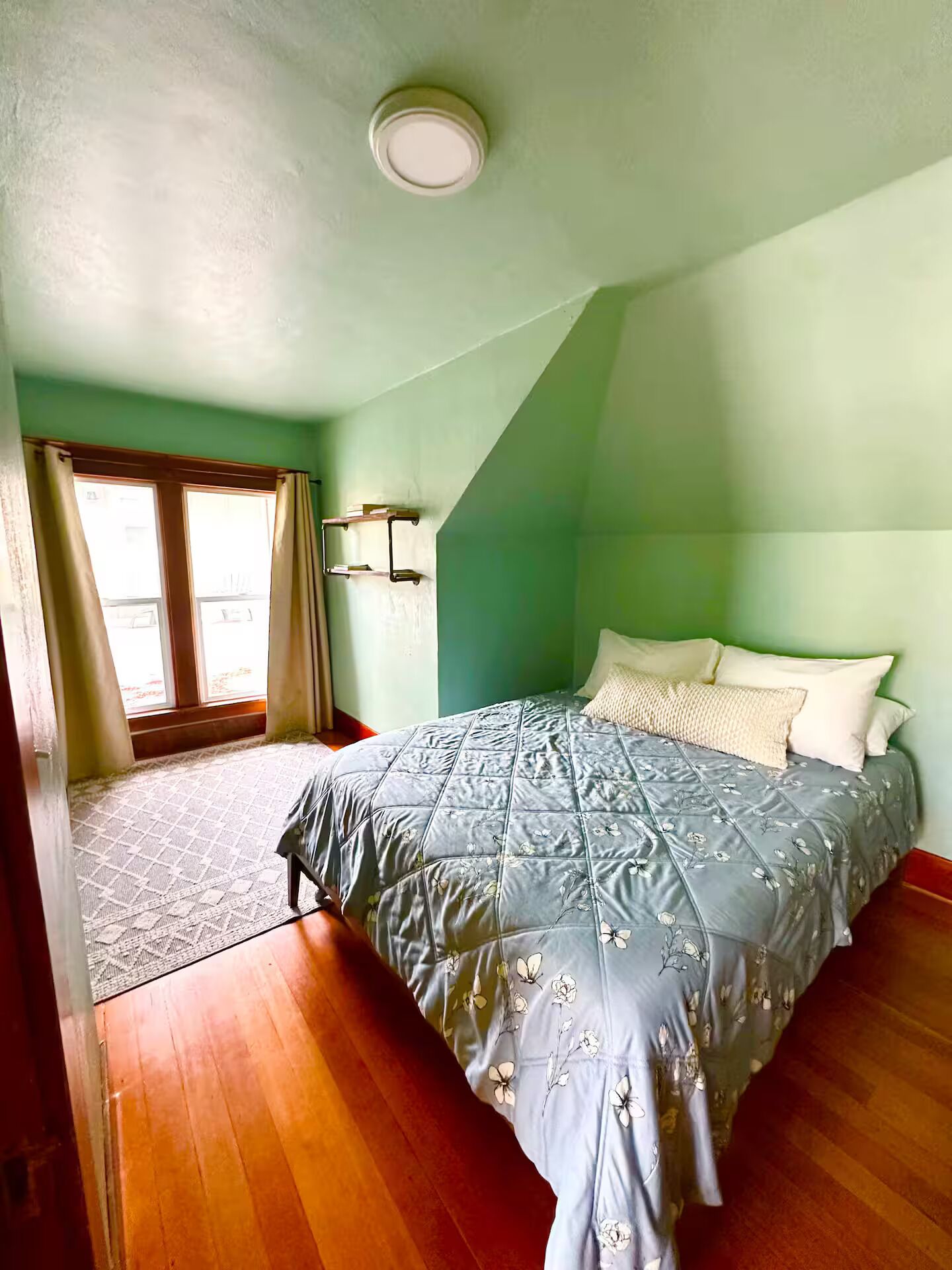 A bedroom with a bed, a window with curtains, a ceiling light, and a wooden floor.